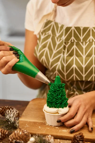 Cropped view of confectioner with pastry bag decorating Christmas tree cupcake on table with spruce cones — Stock Photo