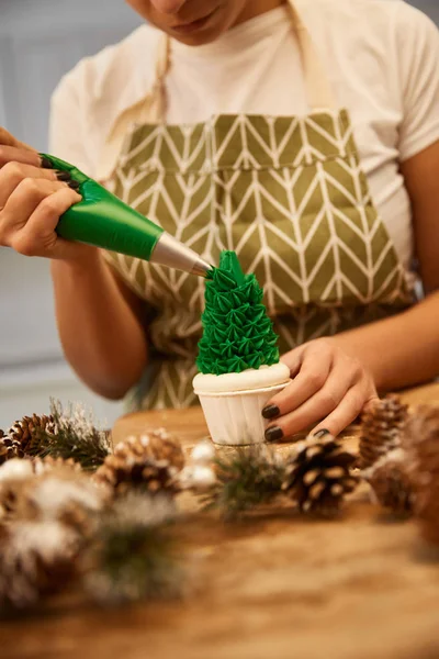 Vista recortada del confitero vertiendo crema verde dulce en el cupcake del árbol de Navidad junto a los conos de abeto en la mesa - foto de stock