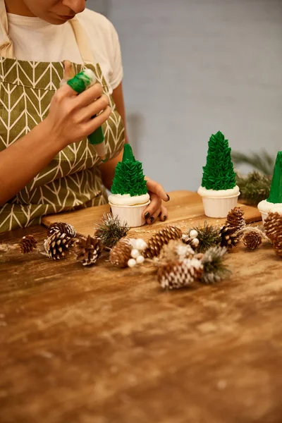 Vista ritagliata del pasticciere decorare cupcakes albero di Natale con crema dolce verde accanto coni di abete rosso sul tavolo — Foto stock