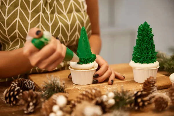 Enfoque selectivo del confitero que trabaja con cupcakes de árbol de Navidad junto a conos de abeto en la mesa, vista recortada - foto de stock