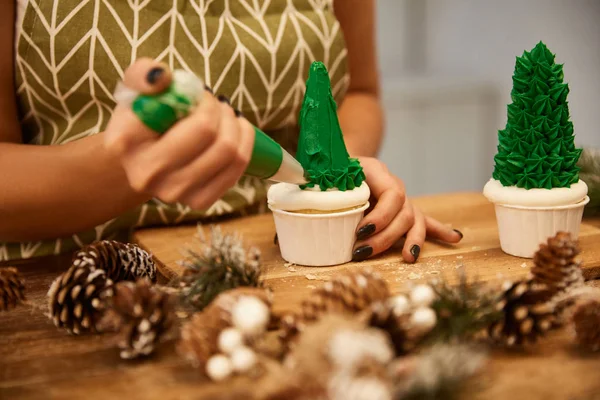 Vista recortada del confitero haciendo cupcakes de árbol de Navidad con crema verde con conos de abeto en la mesa - foto de stock