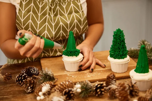 Vista recortada del confitero haciendo cupcakes de árbol de Navidad con crema dulce verde junto a conos de abeto en la mesa - foto de stock