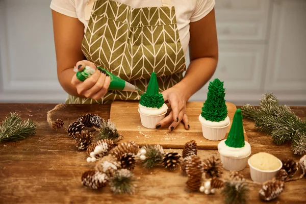 Vista recortada del confitero haciendo cupcakes de árbol de Navidad con crema junto a ramas de abeto y conos en la mesa - foto de stock
