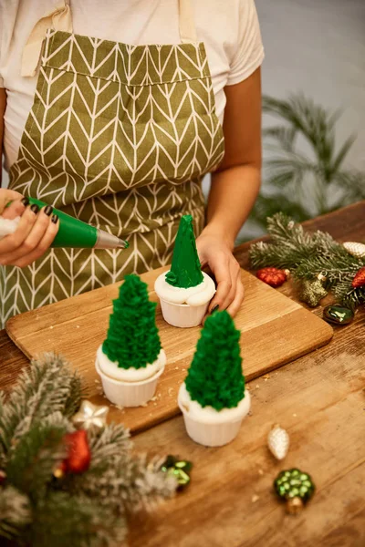 Cropped view of confectioner decorating Christmas tree cupcakes with decorations and spruce branches on table — Stock Photo