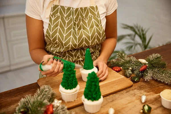 Vista ritagliata del pasticciere che lavora con i cupcake dell'albero di Natale accanto alle decorazioni natalizie e ai rami di abete rosso sulla tavola — Foto stock