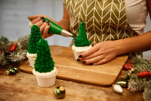 Vista cortada de confeiteiro fazendo cupcakes de árvore de Natal ao lado de ramos de abeto e bolas de Natal na mesa — Fotografia de Stock