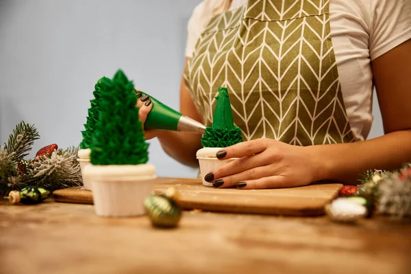 Zugeschnittene Ansicht eines Konditors, der Weihnachtsbaum-Cupcakes neben Tannenzweigen und Weihnachtskugeln auf dem Tisch dekoriert — Stockfoto