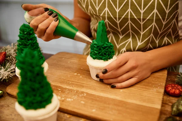 Vue recadrée du confiseur ajoutant de la crème sucrée sur les cupcakes de sapin de Noël avec des branches de pin et des boules de Noël sur la table — Photo de stock