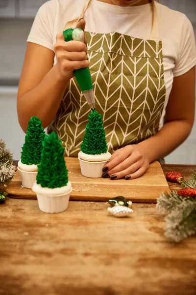 Vue recadrée de confiseur ajoutant de la crème sur les cupcakes de sapin de Noël avec des branches de pin et décor de Noël sur la table — Photo de stock