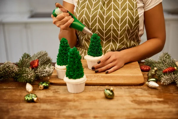 Zugeschnittene Ansicht eines Konditors, der Weihnachtsbaum-Cupcakes neben Fichtenzweigen und Weihnachtsdekor auf dem Tisch herstellt — Stockfoto