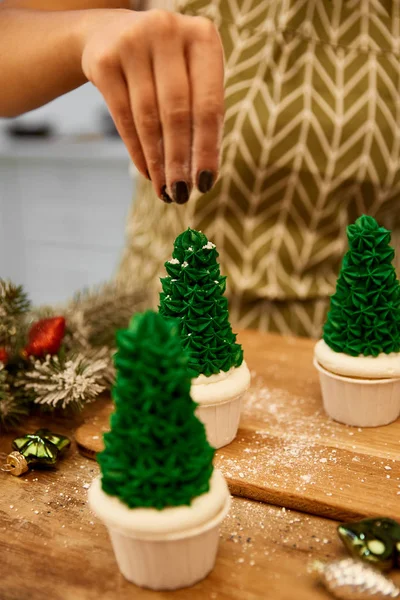 Vista cortada da decoração de aspersão de confeiteiro em cupcakes de árvore de Natal ao lado de bolas de Natal e ramo de abeto na mesa — Fotografia de Stock