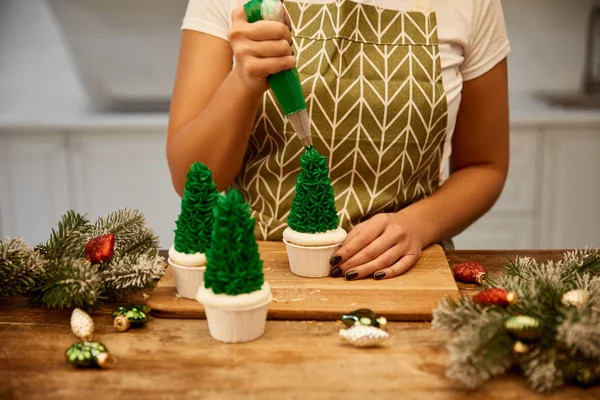 Vista recortada del pastelero con bolsa de pastelería añadiendo crema en cupcakes de árbol de Navidad con ramas de pino en la mesa - foto de stock