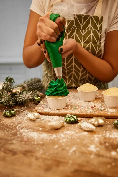 Vue recadrée de confiseur faisant des cupcakes avec de la crème verte à côté de la décoration de Noël et branche de pin sur la table — Photo de stock