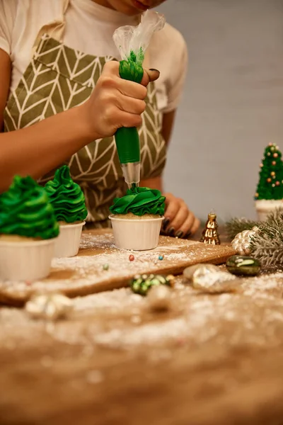 Vista ritagliata del pasticciere che lavora con panna e cupcake accanto a bagattelle e ramo di pino sulla tavola — Foto stock