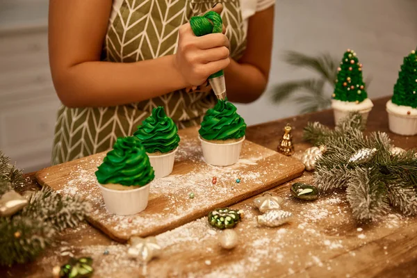 Zugeschnittene Ansicht des Konditors, der Weihnachtsbaum-Cupcakes neben Fichtenzweigen und Weihnachtsschmuck auf dem Tisch dekoriert — Stockfoto