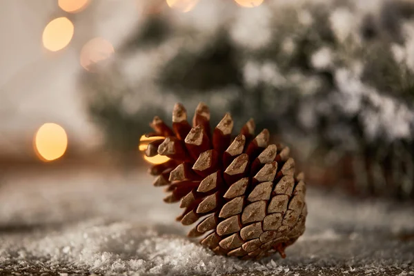 Close up of pine cone on wooden table with spruce branches in snow and christmas lights bokeh — Stock Photo