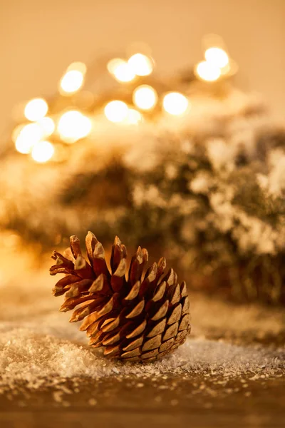 Tannenzapfen auf Holztisch mit Fichtenzweigen im Schnee und Weihnachtsbeleuchtung Bokeh — Stockfoto