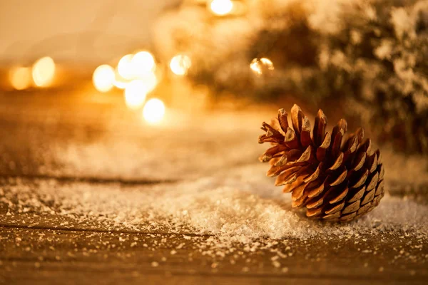 Декоративный сосновый конус на деревянном столе с еловыми ветвями в снегу и рождественские огни боке — стоковое фото