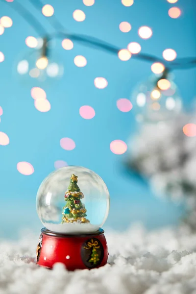 Sapin de Noël en boule de neige debout sur bleu avec neige et lumières floues — Photo de stock