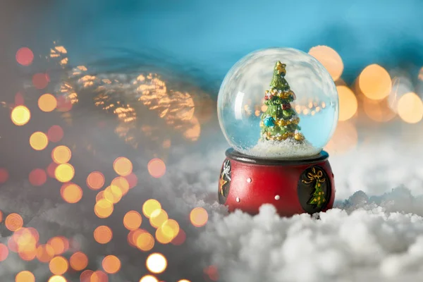 Sapin de Noël en boule de neige debout sur bleu avec neige et lumières floues — Photo de stock