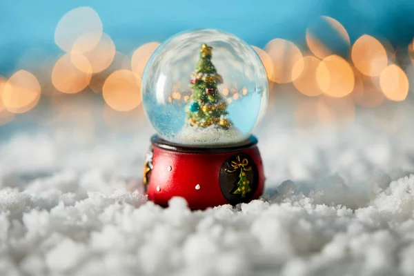 Boule de neige avec sapin de Noël debout sur bleu avec neige et lumières floues — Photo de stock