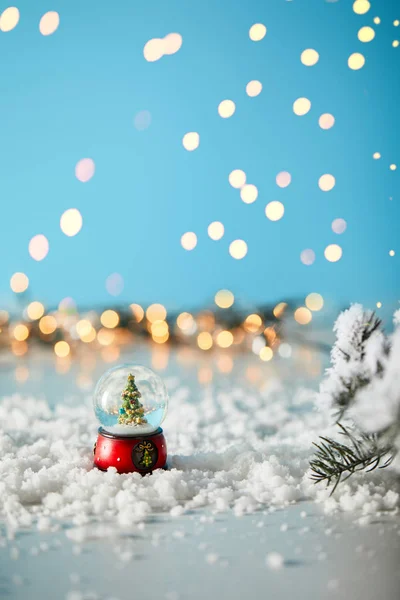 Petit sapin de Noël en boule de neige debout sur bleu avec des branches d'épinette dans la neige et des lumières floues — Photo de stock