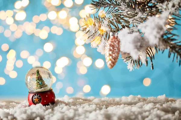 Sapin de Noël en boule de neige debout sur bleu avec des branches d'épinette dans la neige et des lumières bokeh — Stock Photo