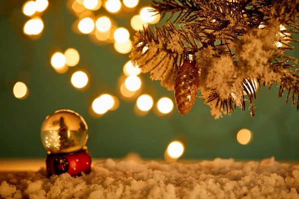 Bola de nieve con árbol de Navidad de pie en la nieve con ramas de abeto, bola de Navidad y luces borrosas por la noche - foto de stock