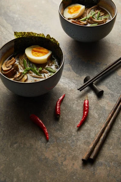 Traditional spicy ramen in bowls near chopsticks and vegetables on stone surface — Stock Photo