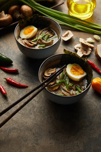 Traditional spicy ramen in bowls near chopsticks and vegetables on stone surface — Stock Photo