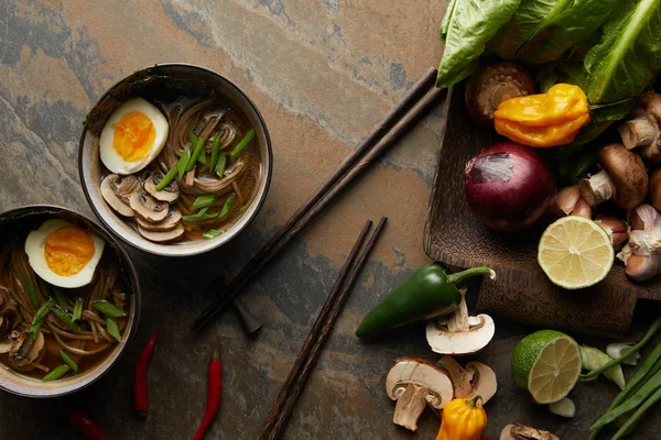 Вид сверху на традиционный пряный рамен в мисках возле палочек для еды и овощей на каменной поверхности — стоковое фото