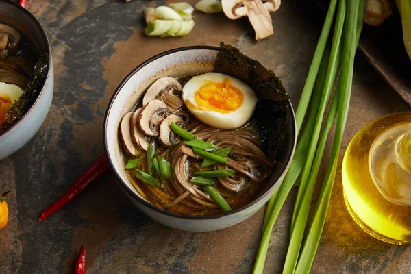 Traditional spicy ramen in bowl near vegetables and oil on stone surface — Stock Photo