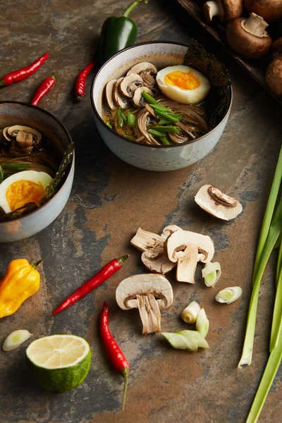 Traditional spicy ramen in bowls near vegetables on stone surface — Stock Photo
