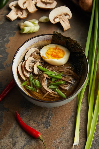 Traditional spicy ramen in bowl near vegetables on stone surface — Stock Photo