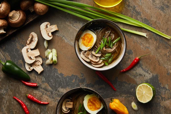 Top view of traditional spicy ramen in bowls near vegetables  and oil on stone surface — Stock Photo