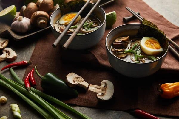 Traditional spicy ramen in bowls with chopsticks and vegetables on brown napkin on stone surface — Stock Photo