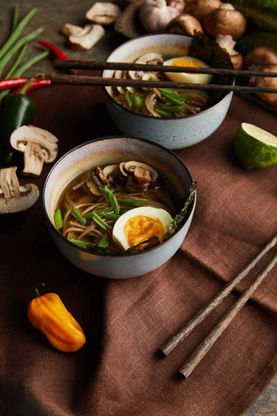 Ramen picante tradicional em tigelas com pauzinhos e legumes em guardanapo marrom na superfície de pedra — Fotografia de Stock