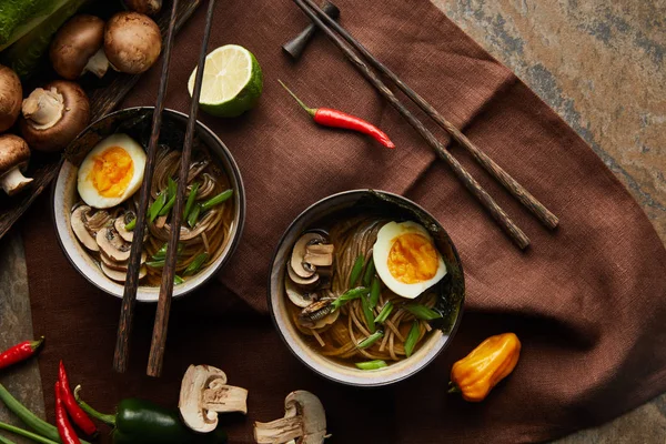 Vue de dessus des ramen épicés traditionnels dans des bols avec baguettes et légumes sur une serviette brune sur la surface de la pierre — Photo de stock