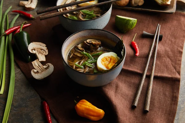 Traditional spicy ramen in bowls with chopsticks and vegetables on brown napkin on stone surface — Stock Photo