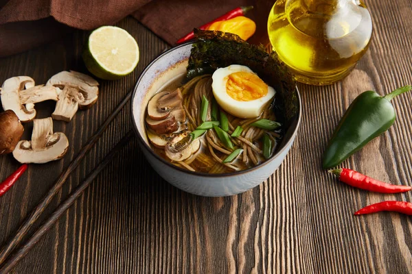 Traditional spicy ramen in bowl with chopsticks and vegetables on wooden table with napkin — Stock Photo