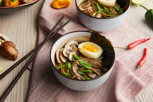 Traditional spicy ramen in bowls with chopsticks and vegetables on wooden table with napkin — Stock Photo