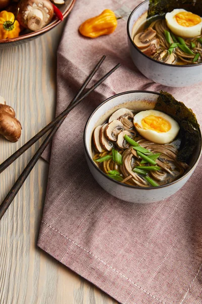 Traditional spicy ramen in bowls with chopsticks and vegetables on wooden table with napkin — Stock Photo
