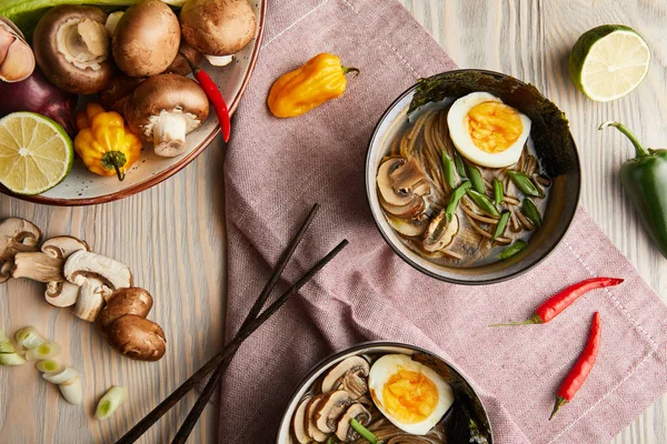 Vista superior do ramen picante tradicional em tigelas com pauzinhos e legumes em mesa de madeira com guardanapo — Fotografia de Stock