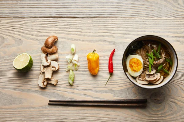 Posa piatta con ramen speziato tradizionale in ciotola vicino bacchette e ingredienti sul tavolo di legno — Foto stock