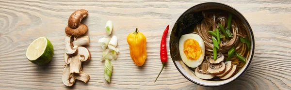 Flat lay with traditional spicy ramen in bowl near chopsticks and ingredients on wooden table, panoramic shot — Stock Photo