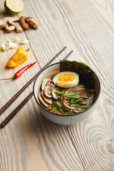 Selective focus of traditional spicy ramen in bowl near chopsticks and ingredients on wooden table — Stock Photo
