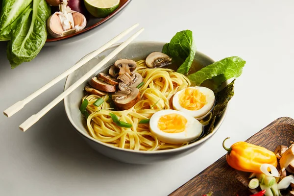 Traditional asian ramen in bowl near chopsticks, vegetables on grey surface — Stock Photo