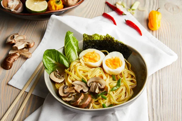 Traditional asian ramen in bowl near chopsticks, napkin and vegetables on wooden table — Stock Photo