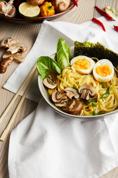 Traditional asian ramen in bowl near chopsticks, napkin and vegetables on wooden table — Stock Photo