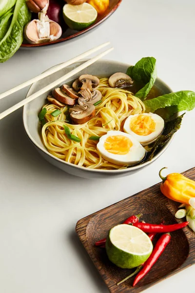 Traditional asian ramen in bowl near chopsticks, vegetables on grey surface — Stock Photo